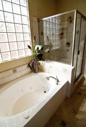 Modern bathtub and shower combination in well-lit bathroom featuring glass block window, decorative vase with flowers, and tiled surfaces, relevant to tub and shower replacement considerations.