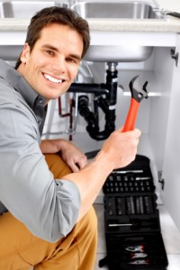 Smiling plumber holding a wrench under a kitchen sink, showcasing plumbing tools and pipes, relevant to garbage disposal installation and repair services.