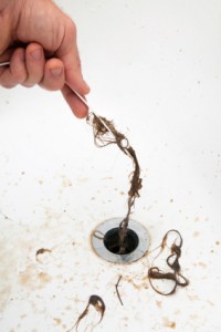 Hand removing hair and debris from a clogged drain, illustrating the importance of annual drain cleaning and maintenance services for preventing plumbing issues.