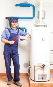 Plumber smiling next to a water heater, holding tools, in a residential setting, emphasizing water heater maintenance and repair services.