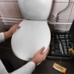 Person performing maintenance on a toilet, with plumbing tools and a toolbox visible nearby, emphasizing toilet upkeep to prevent clogs.