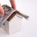 Hand placing cash on a model house, symbolizing the costs of plumbing repairs and the value of professional services.