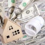 Model house surrounded by money and an energy-efficient light bulb, symbolizing savings and efficiency in residential energy costs.