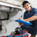 Plumber inspecting under a sink with tools and a clipboard, emphasizing professional plumbing services for drain cleaning and maintenance.