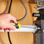 Person using a screwdriver to repair a garbage disposal unit under a kitchen sink, emphasizing plumbing maintenance and repair services.